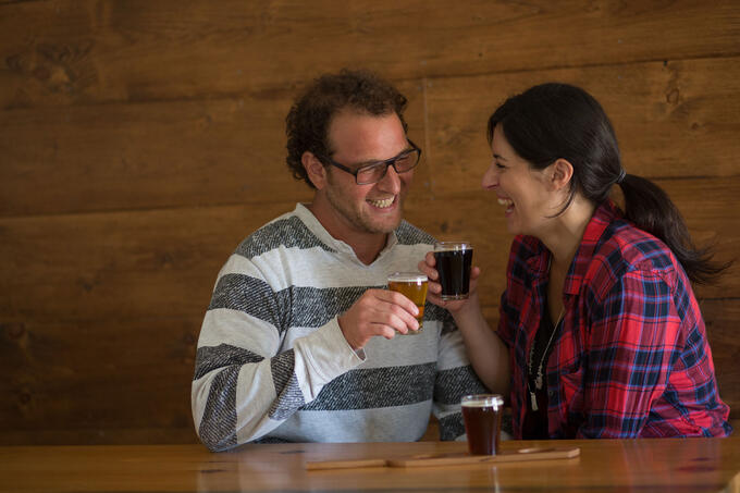 two people sitting at a wooden table enjoying a flight of beer; they are laughing and clinking their drink glasses together